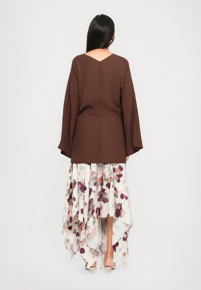 Woman wearing a brown long-sleeve top and an asymmetrical white skirt with purple and orange floral patterns, facing away, on white background.