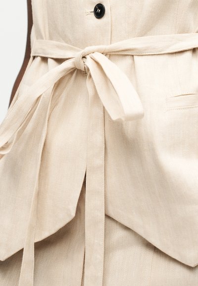 Ceinture en tissu beige nouée à la taille sur un vêtement assorti boutonné avec détail de poche.