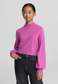Maglione rosa accorciato con colletto a lupetto e maniche a palloncino, abbinato a jeans neri a vita alta con chiusura a bottoni.