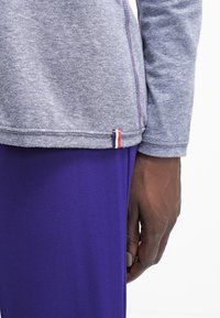 Grå långärmad topp med kontrasterande röd söm och en liten röd, vit och blå etikett i fållen, kombinerad med enfärgade lila byxor.