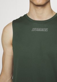 Ciemnozielona bezrękawnik sportowy wykonany z teksturowanej tkaniny, z okrągłym dekoltem i kontrastowym białym logo "HUMMEL" na piersi.