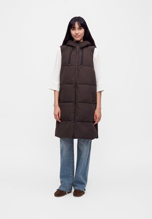 Vrouw met een lange bruine gewatteerde bodywarmer met capuchon, witte blouse met lange mouwen, blauwe spijkerbroek en bruine schoenen, staand tegen een effen achtergrond.