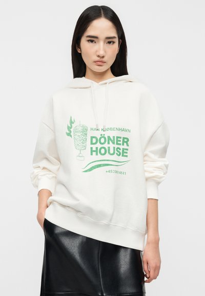 Νεαρή γυναίκα που φοράει λευκό φούτερ με κουκούλα με πράσινο κείμενο "Döner House", γραφικό σουβλακιού και μαύρη δερμάτινη φούστα, στέκεται μπροστά σε ένα απλό φόντο.
