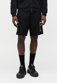 Μαύρα βαμβακερά σορτς με ίσια γραμμή, με λεπτομέρεια λογότυπου στην αριστερή πλευρά, συνδυασμένα με μαύρες κάλτσες και αθλητικά παπούτσια.