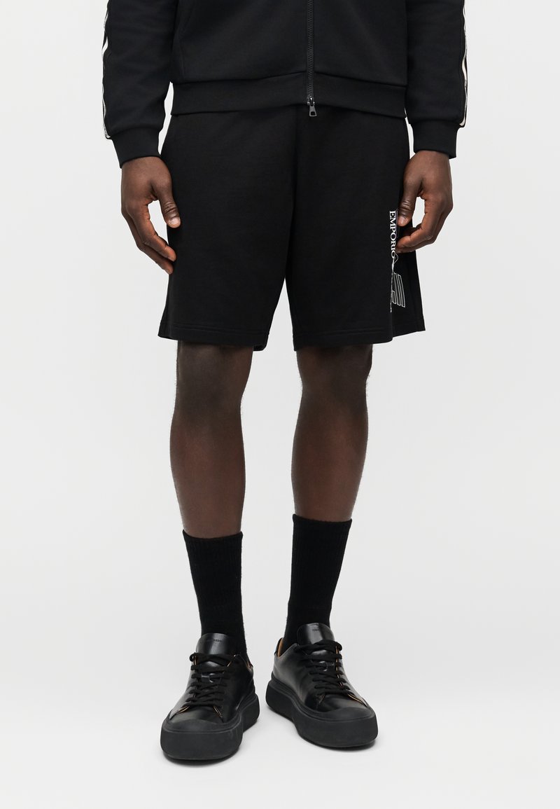 Μαύρα βαμβακερά σορτς με ίσια γραμμή, με λεπτομέρεια λογότυπου στην αριστερή πλευρά, συνδυασμένα με μαύρες κάλτσες και αθλητικά παπούτσια.