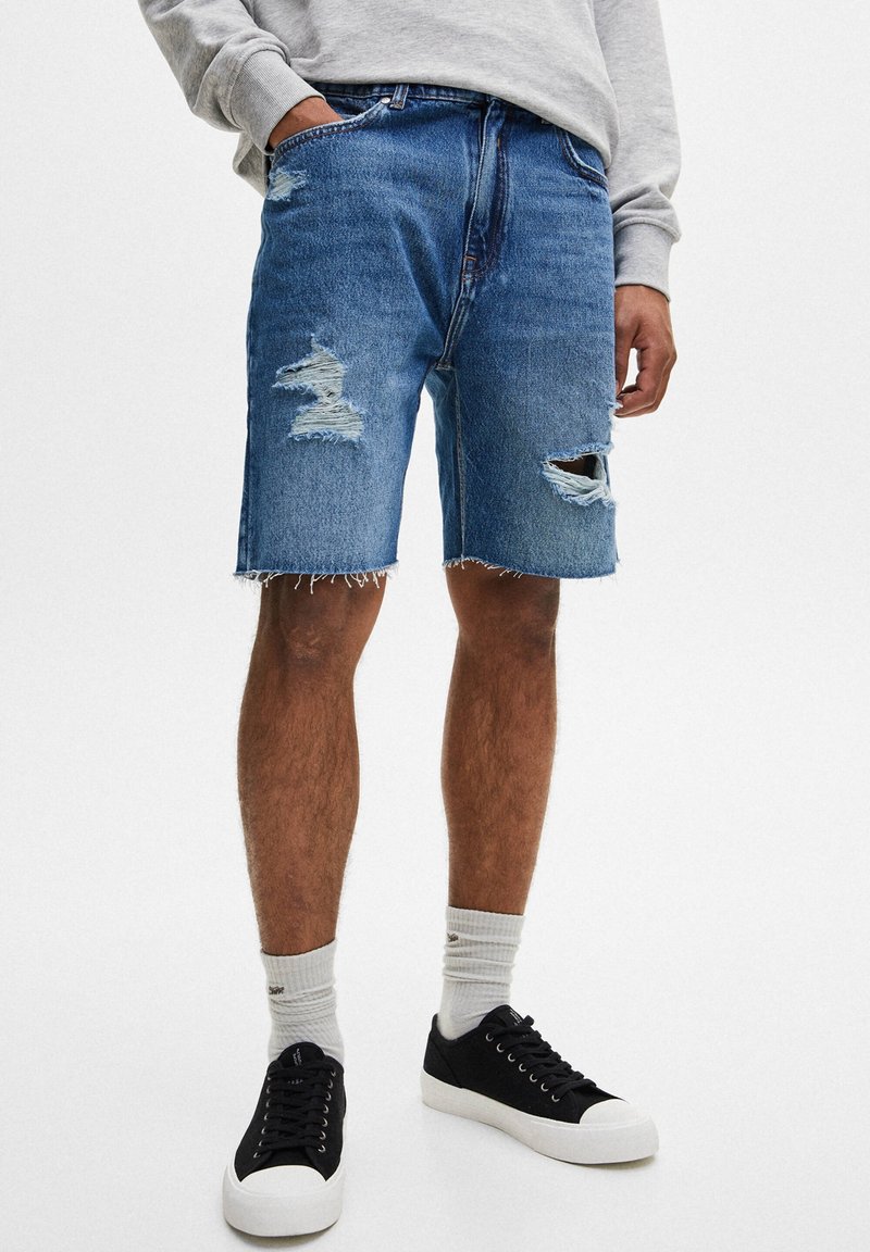 Shorts en denim de bleu moyen avec des patchs déchirés. Présente un ourlet effiloché, cinq poches, et est porté avec des chaussures noires et des chaussettes gris clair.