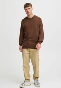 Young man standing in beige pants, brown sweater, and white sneakers against a plain light background.