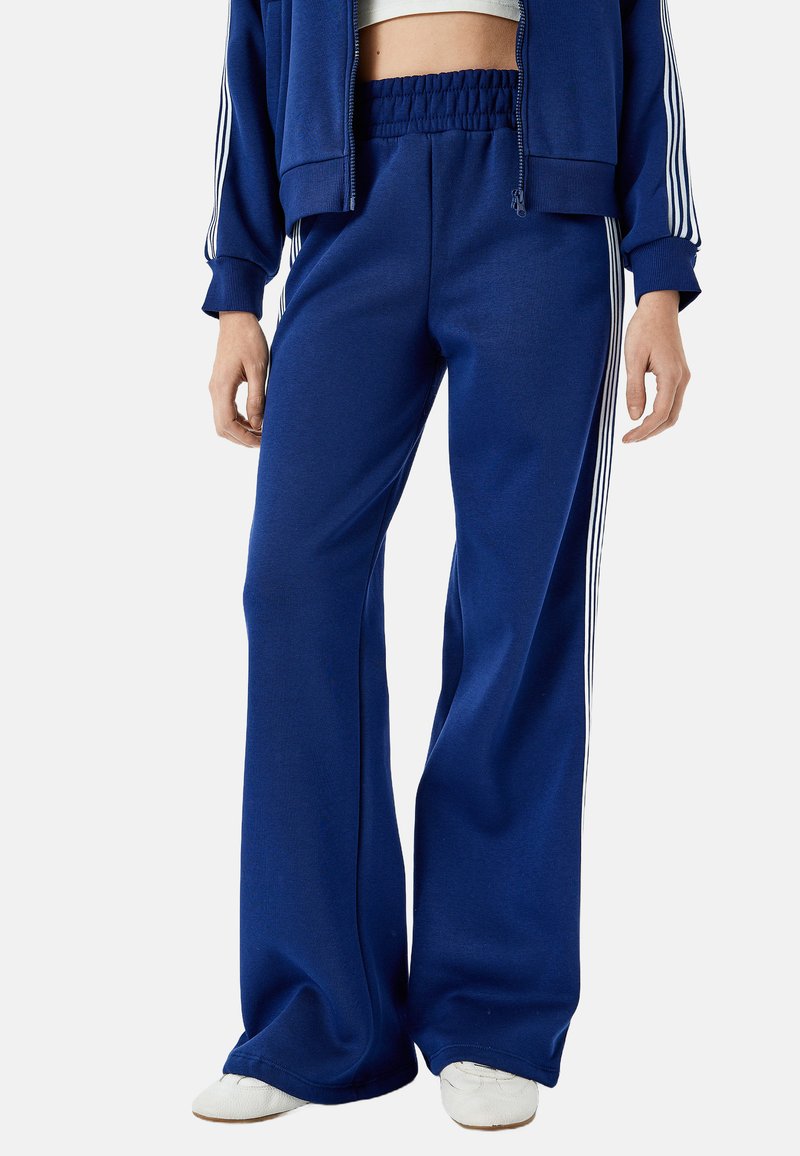 Blå sweatpants i mjukt tyg, med elastisk midja, lös passform och tre vertikala vita ränder längs sidorna.