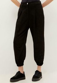 Zwarte, cropped broeken van zachte stof met plooien, een knoopsluiting en elastische boorden; gecombineerd met zwarte sneakers.