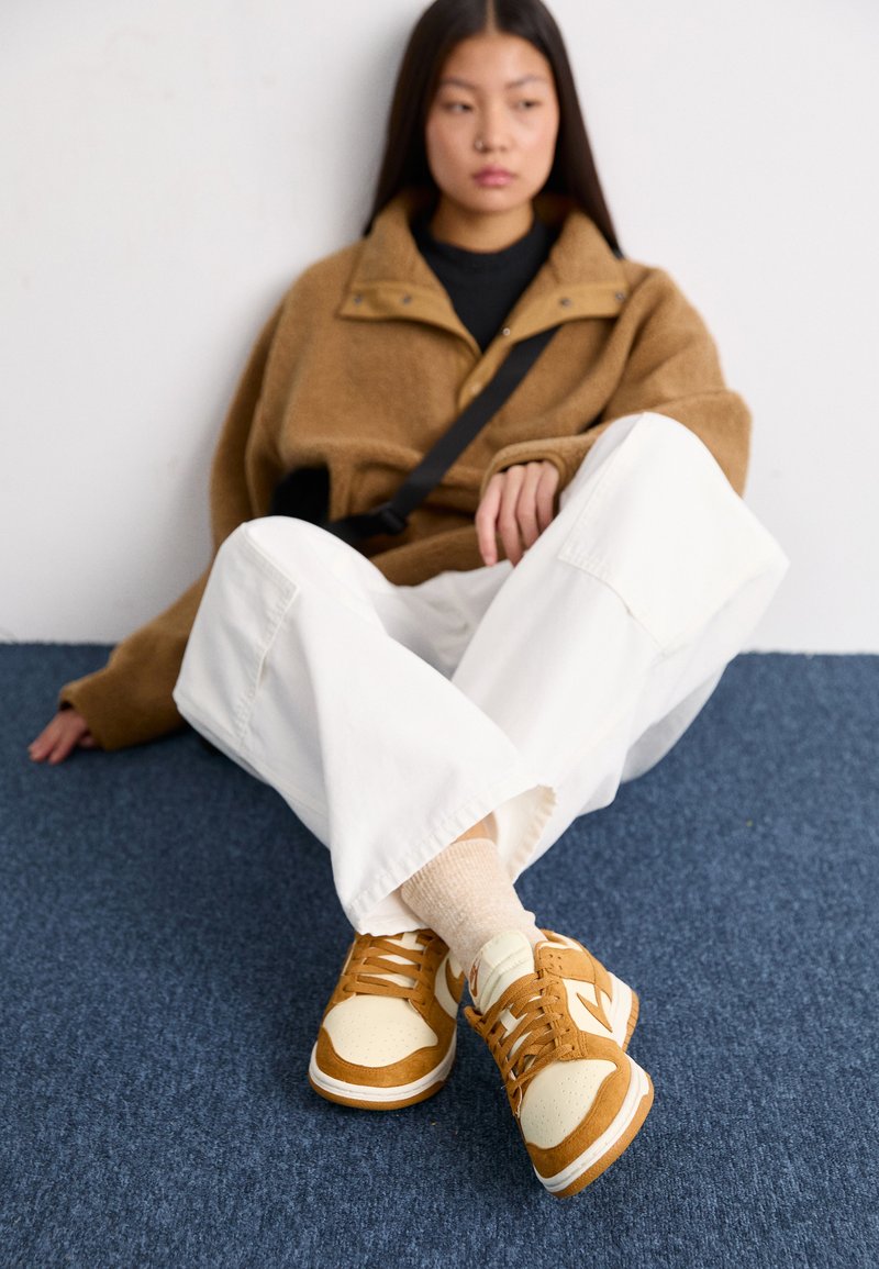 Baskets aux teintes beige et caramel, avec un mélange de cuir et de daim. Des lacets larges, une boîte à orteils perforée et une semelle blanche sont des détails marquants.