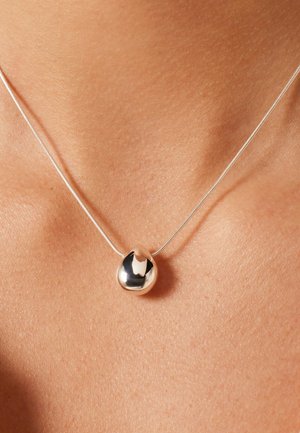 Glanzende, onregelmatige druppelvormige zilveren hanger aan een dunne zilveren ketting, die rust op blote huid in het midden van de borst.