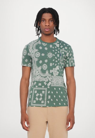 T-shirt vert à manches courtes avec des motifs paisley et floraux blancs, encolure ronde et tissu texturé. Porté avec un pantalon beige.