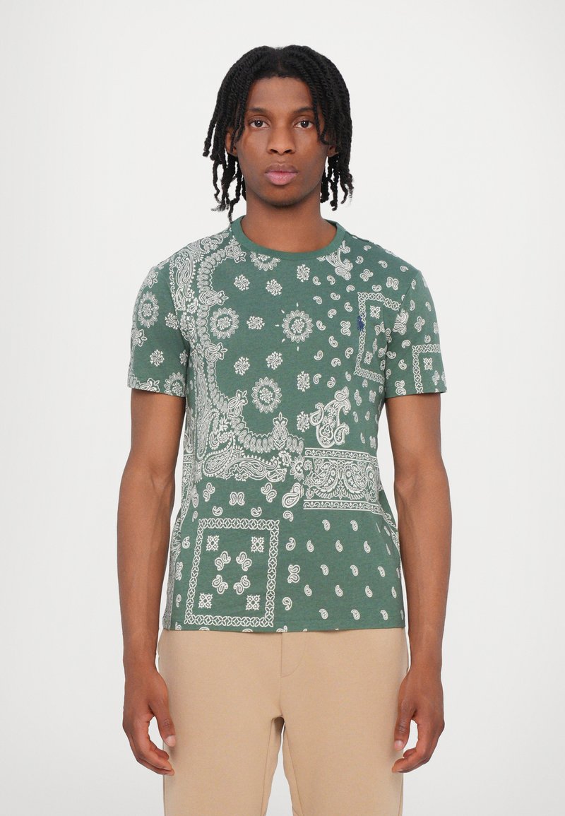Groene t-shirt met korte mouwen, versierd met witte paisley- en bloemenpatronen, ronde halslijn en een structuurstof. Draagt met tan kleurige broek.