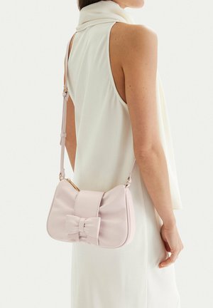 Kobieta w białej bluzce bez rękawów nosi bladoróżową torebkę na ramię z kokardą po boku.