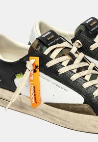 Sneakers a montante basso con pannelli neri, bianchi e mimetici, lacci beige, etichetta arancione "Crime London" e suola in gomma beige consumata.
