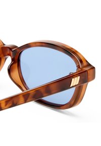 Κοντινή λήψη του μπράτσου και του σκελετού γυαλιών ηλίου με χελωνόκρανο, με φακούς ανοιχτού μπλε χρώματος και μικρή χρυσή διακοσμητική λεπτομέρεια.