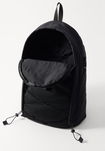 Sac à dos noir avec un extérieur lisse, une poche en maille à l'avant et une fermeture à cordon réglable à la base. Le rabat ouvert révèle l'intérieur.