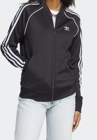 Person som bär en svart Adidas zip-up-jacka med vita ränder på ärmarna och logotyp, tillsammans med ljusblå jeans och kedjehalsband.