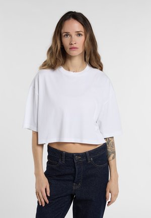 Kvinna med långt hår som bär en lös vit kort t-shirt och mörkblå jeans, som visar en blommig tatuering på vänster underarm.