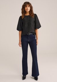 Žena má na sebe čierny textúrovaný top s nariasenými rukávmi, tmavomodré nohavice s vysokým pásom a rozšírenými nohavicami, čierny opasok a čierne špicaté topánky.