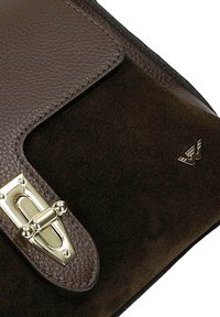 Patta della borsa in pelle scamosciata marrone scuro e pelle martellata con chiusura a girello color oro e piccolo emblema metallico sulla superficie scamosciata.