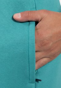 Zielona tkanina bawełniana z domieszką, z ukrytą boczną kieszenią i dyskretnym czarnym sznurkiem. Gładka faktura z prążkowanymi detalami.