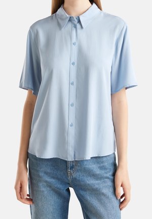 Chemise à manches courtes bleu clair boutonnée portée avec un jean bleu sur une personne debout montrée du cou aux hanches.