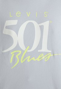 Logo Levi's 501 Blues imprimé sur un tissu bleu clair avec un texte blanc et jaune néon en polices mixtes.