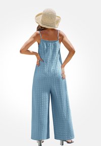 Lichtblauwe jumpsuit met wijde benen, gestructureerde stof, elastische ruches aan de halslijn en dunne bandjes. Gecombineerd met een strooien zonnehoed.