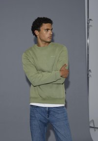 Felpa verde con scollo a girocollo e logo "blend" ricamato, abbinata a jeans blu. Sfondo grigio, con illuminazione da studio.