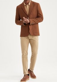 Bruine blazer met textuur en twee opgestikte zakken, lichtbeige overhemd, beige slim-fit broek en bruine sneakers met zwarte accenten.