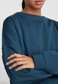Round-neck sweatshirt in deep teal, made from soft fabric. Features dropped shoulders and a relaxed fit. Hands with light-colored nails rest on the arm.