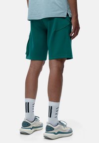 Groene sportshorts met een ontspannen pasvorm, voorzien van zijritszakken. Draagt witte sokken en lichtblauwe sneakers.