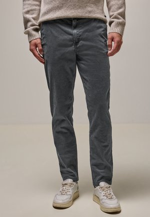 Mann trägt graue, strukturierte Hose, beigen Pullover und weiße Sneaker, steht vor einfachem Hintergrund.