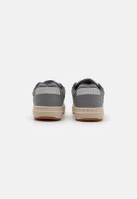 Sive kožne tenisice s niskim profilom, teksturiranim stražnjim dijelom i svijetlosmešnatim potplatima. Sadrži "CONVERSE" oznaku na peti.