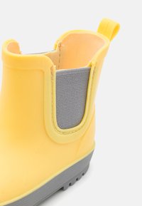 Dzeltenas gumijas zābaks ar pelēku elastīgu sānu paneļu, gludu virsmu un strukturētu pelēku zoli labākai saķerei.