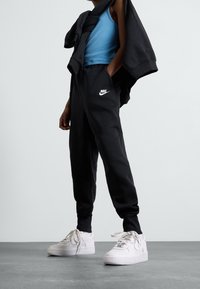 Schwarze Jogginghosen mit einem weißen Nike-Logo, kombiniert mit einem blauen Tanktop und einem schwarzen Sweatshirt, das über die Schultern drapiert ist; weiße Sneaker mit Schnürsenkeln.