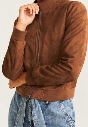 Persona que lleva una chaqueta de ante marrón con cremallera, puños y cuello acanalados, combinada con unos vaqueros de tiro alto azul claro con cinturón anudado.