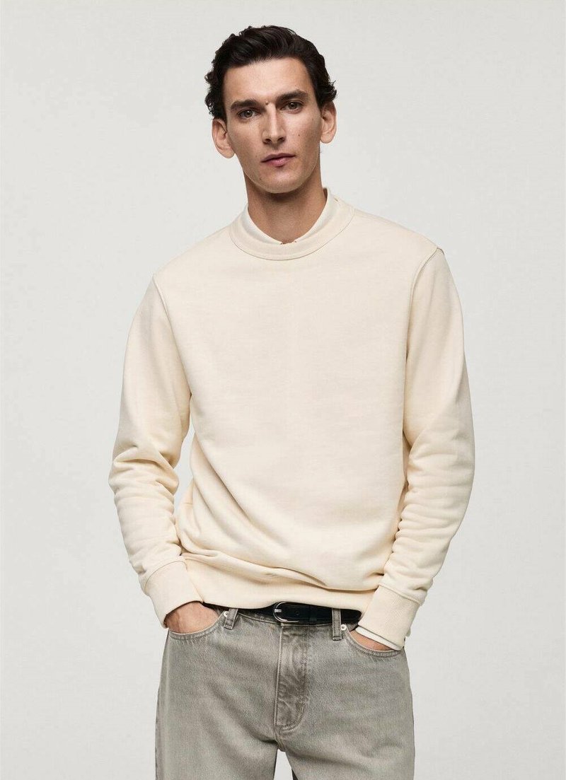 Cream-colored sweatshirt with a round neck, ribbed cuffs, and hem. Worn with light grey jeans. Smooth texture, minimal design.