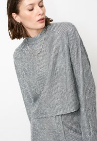 Zilveren, gemeleerde crop top met lange mouwen en een hoge halslijn, voorzien van een subtiel gestreept patroon. Het model draagt een ketting.
