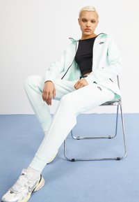 Mintgroene Nike trainingspak, zachte stof, rits hoodie, slim-fit broek, zwart logo op de borst en bovenbeen, gecombineerd met zilveren en witte sneakers.