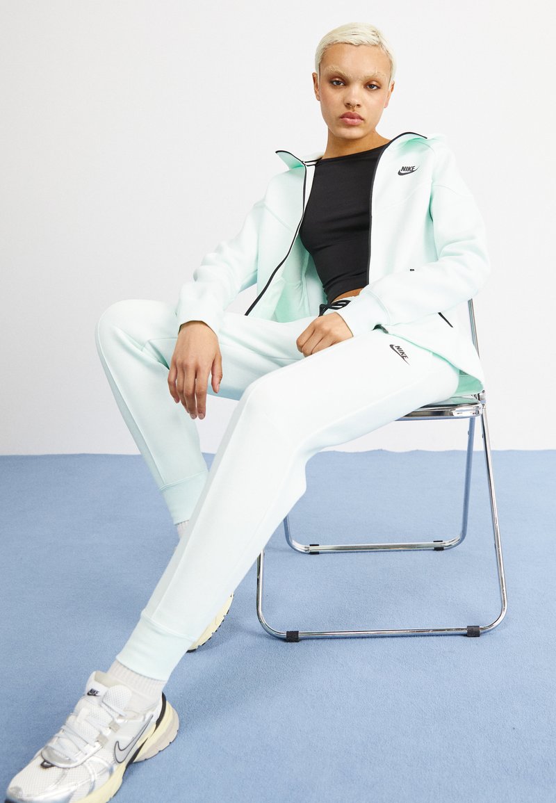 Tuta Nike verde menta, tessuto morbido, felpa con zip, pantaloni aderenti, logo nero su petto e coscia, abbinato a scarpe da ginnastica argentate e bianche.