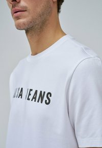 Camiseta de algodón blanca con un audaz texto en negro que dice "ALISA JEANS". Manga corta y cuello redondo. Textura suave con un ajuste relajado.