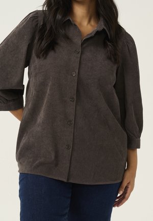 Femme portant une chemise boutonnée en velours côtelé marron foncé à manches bouffantes et un jean bleu, debout devant un fond uni.