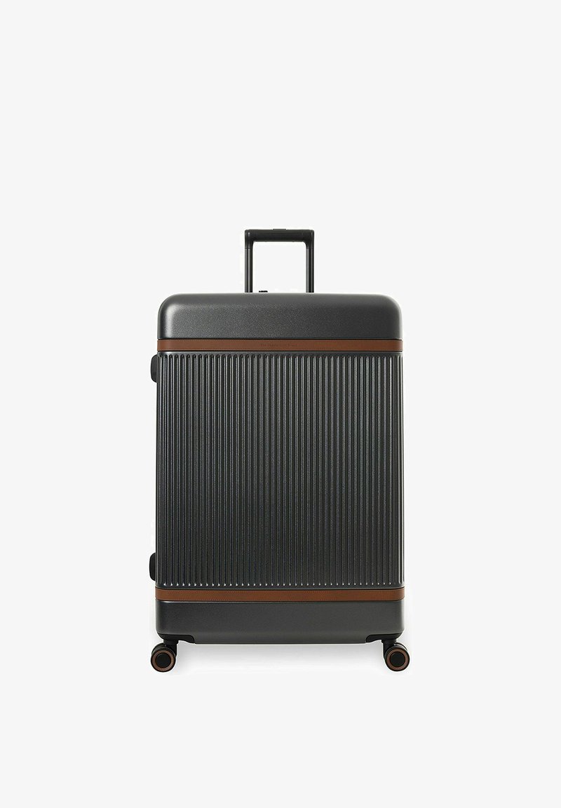 Valise rigide noire avec des rainures verticales, accents horizontaux marron, poignée rétractable déployée, et quatre roues multidirectionnelles sur fond blanc.