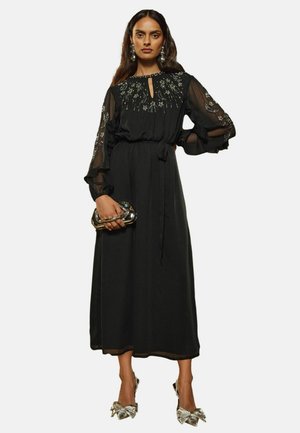 Rochie neagră cu mâneci lungi transparente, guler tip cheie, broderie florală, talie strânsă și fustă fluidă. Asortată cu o poșetă metalică și tocuri.