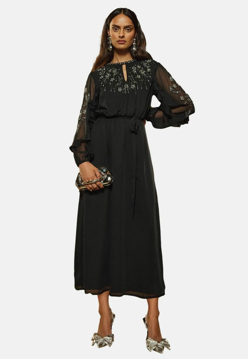 Čierne šaty s priesvitnými dlhými rukávmi, výstrihom do tvaru kľúčovej dierky, kvetinovou výšivkou, úzkym pásom a splývavou sukňou. V kombinácii s metalickou kabelkou a lodičkami.