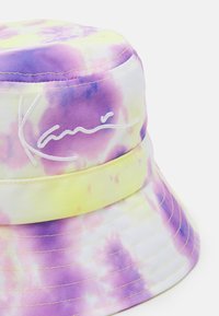 Bøttehatt med tie-dye mønster i lilla og gule farger. Laget av tekstil med sømte detaljer og brodert merkenavn.