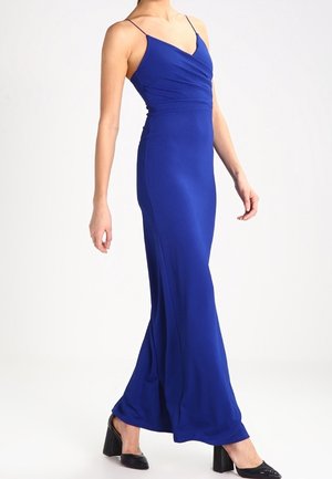 Frau trägt ein bodenlanges, königsblaues, ärmelloses Kleid mit dünnen Trägern und schwarzen Schuhen mit Blockabsatz, steht vor einem weißen Hintergrund.