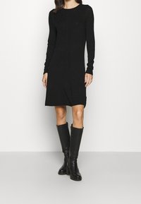 Schwarzes Langarmkleid mit einer figurbetonten Silhouette, geripptem Textur an den Schultern und einem gewellten Saum. Kombiniert mit knielangen schwarzen Stiefeln.